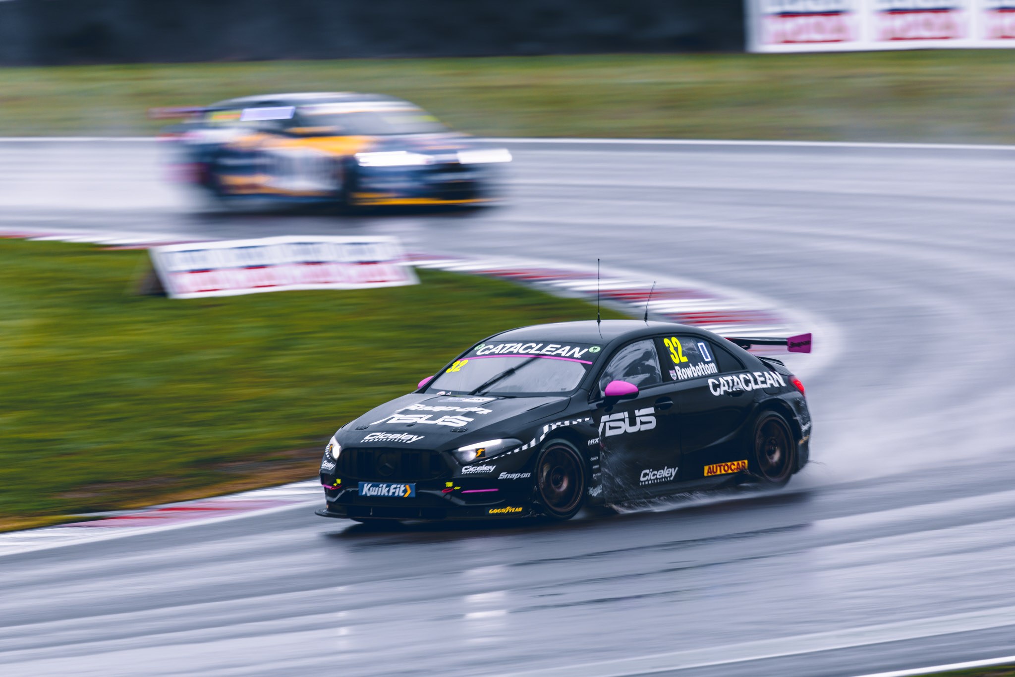 Black number 32 touring car with pink mirrors rounding a wet corner, with another car behind.