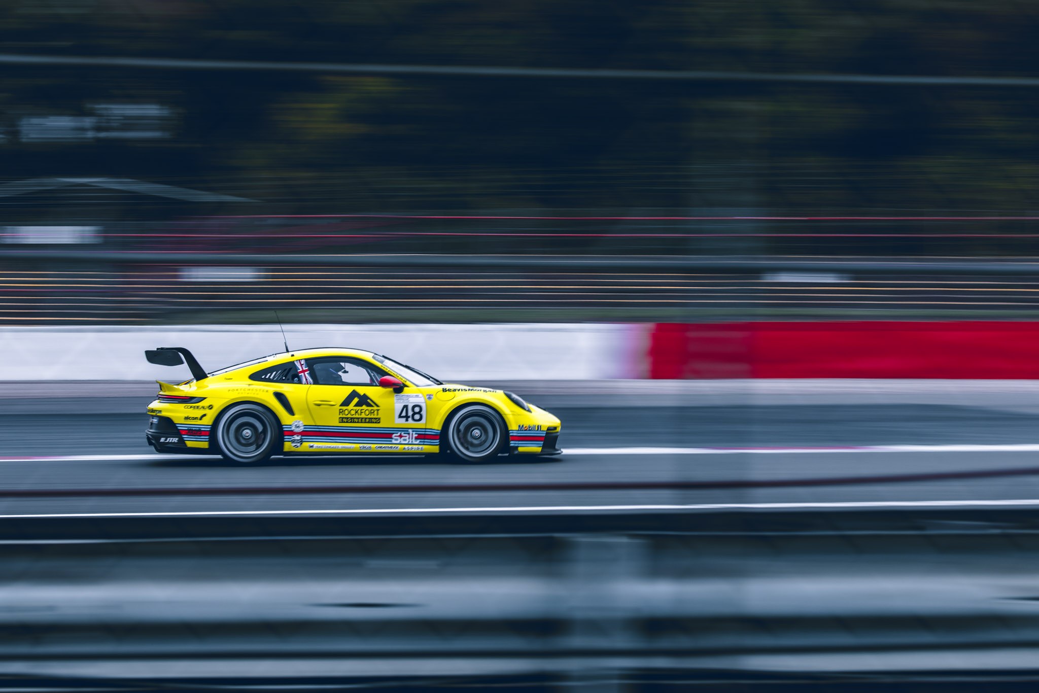 Yellow number 48 Porsche racing car photographed side-on, with the barriers and background streaked by motion.