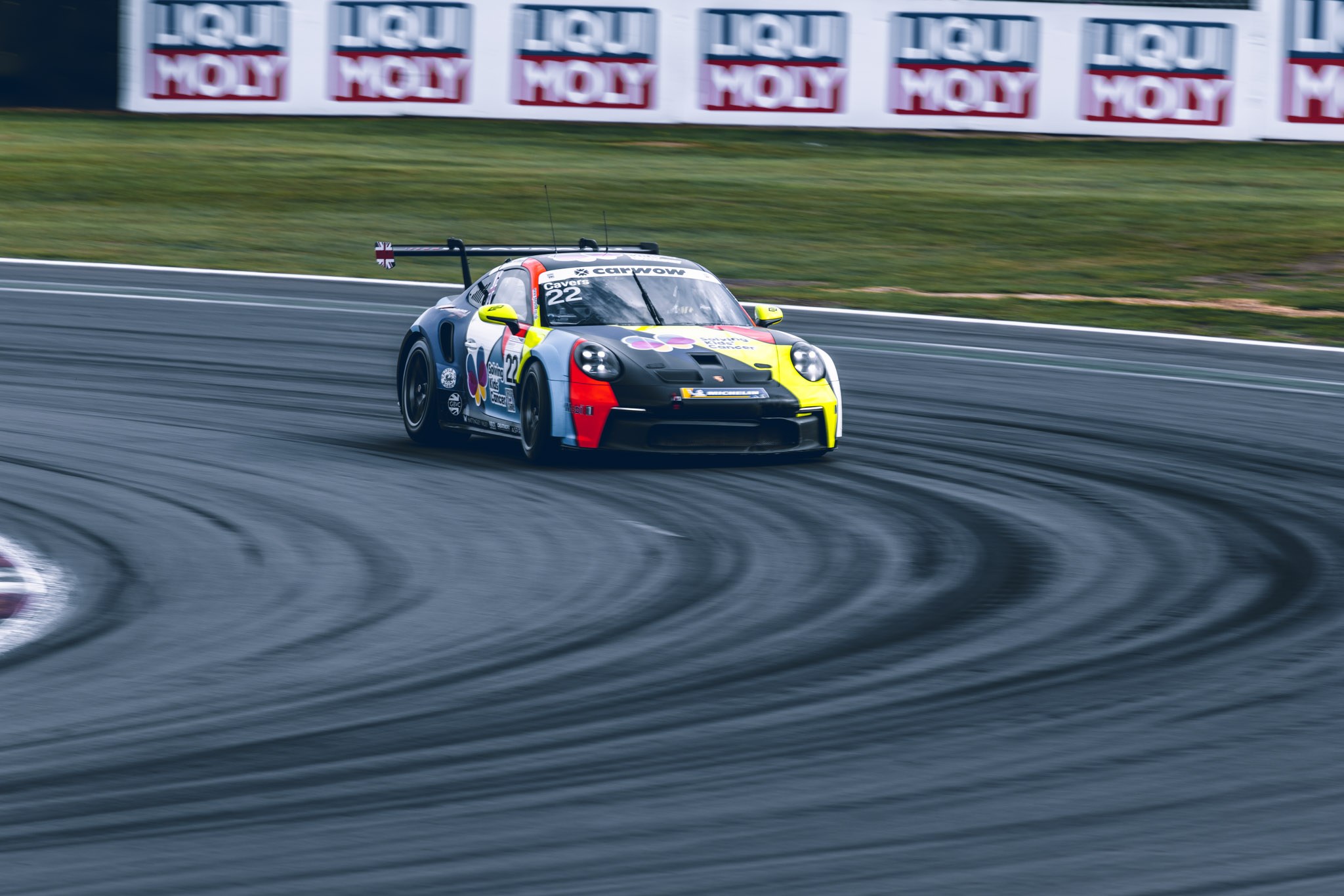 Porsche racing car with yellow and red accents turning through a sweeping corner, with tyre marks across the tarmac.