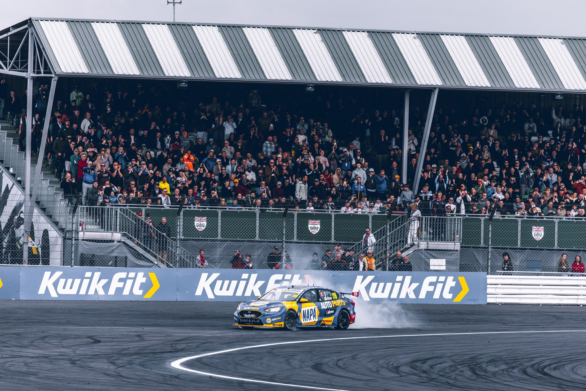 Blue and yellow NAPA touring car passing a grandstand filled with spectators.